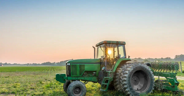 A green tractor parked in the middle of a green field during the day. The sun is high in the sky behind the tractor.